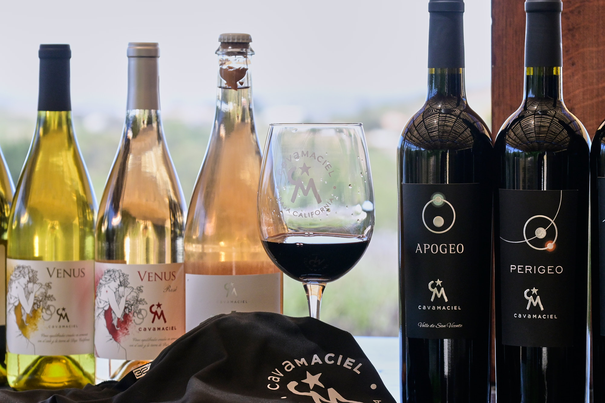 Botella de vino tinto Cava Maciel sobre mesa de madera, acompañada de copa servida y fondo desenfocado de viñedos