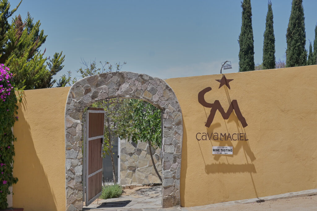 Entrada principal de Cava Maciel con arco de piedra y muros de adobe, enmarcada por viñedos y cielo despejado del Valle de Guadalupe.